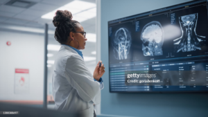 A neurosurgeon standing in a hospital looking at a monitor displaying scan data of a human head and brain